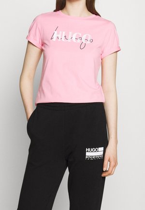 Kobieta w różowym T-shircie z napisem "HUGO" i czarnych spodniach z białym logo "HUGO" na lewym udzie, stojąca z jedną ręką w kieszeni.