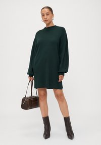 Sötétzöld kötött ruha magas nyakkivágással és lecsúszott vállvarratokkal. Barna táskával és bokáig érő, hegyes orrú csizmával párosítva.