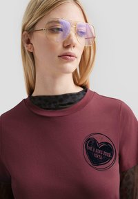 Maronenfarbenes Baumwoll-T-Shirt mit einem runden, dunklen Grafikdesign und Text in Herzform. Darunter getragen ein schwarzes, gemustertes Langarmoberteil.