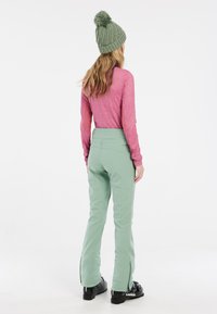 Pantaloni da sci verde menta con vita alta e cerniere laterali, abbinati a una maglietta a maniche lunghe rosa e a un berretto in maglia verde con pompon.