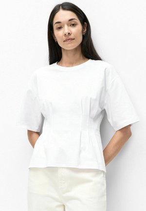 Vrouw met lang donker haar die een simpel wit T-shirt met korte mouwen en lichte broek draagt, staand tegen een witte achtergrond.