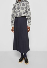 Šedá mikina s opakujícím se potiskem emblému a tmavě modrá plisovaná midi sukně. Doplňky zahrnují černé mokasíny a tmavě modré ponožky.