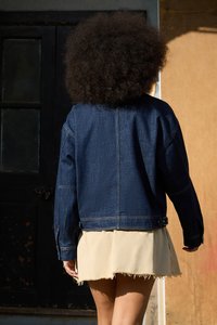 Mørkeblå denimjakke med avslappet passform, med synlige sømmer og lange ermer, brukt over et lysbeige skjørt med frynsete kanter.