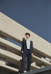 Completo blu navy con giacca doppiopetto, camicia bianca con colletto, in piedi su una struttura di cemento sotto un cielo blu limpido.