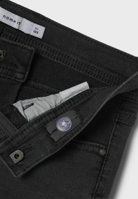 Pantalon en denim noir avec une texture lisse, doté d'un bouton en métal argenté et d'une fermeture éclair. Poche intérieure avec une doublure gris clair.