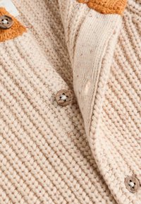 Virkat kofta i beige med en orange detalj. Har texturerat tyg, träknappar och en lös krage.