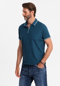 Tyrkysové polo tričko z ribkované tkaniny, má límec, krátké rukávy a bílé lemy kolem límce a rukávů. Nosené s modrými džíny.