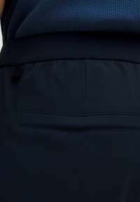 Primer plano de la cinturilla de unos pantalones azul marino con un único bolsillo trasero tipo ribete y una camiseta de azul oscuro texturizada metida por dentro.