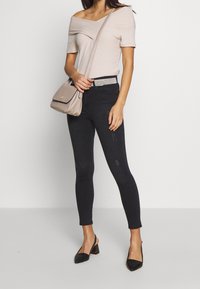 Top beige con hombros descubiertos, jeans ajustados oscuros con detalles desgastados, tacones slingback negros y un bolso ligero con solapa.