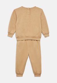 Σετ αθλητικής φόρμας δύο τεμαχίων από απαλό, μπεζ ύφασμα. Το πάνω μέρος έχει στρογγυλή λαιμόκοψη και ριμπ μανσέτες· το παντελόνι διαθέτει ελαστικές μπατζάκια.