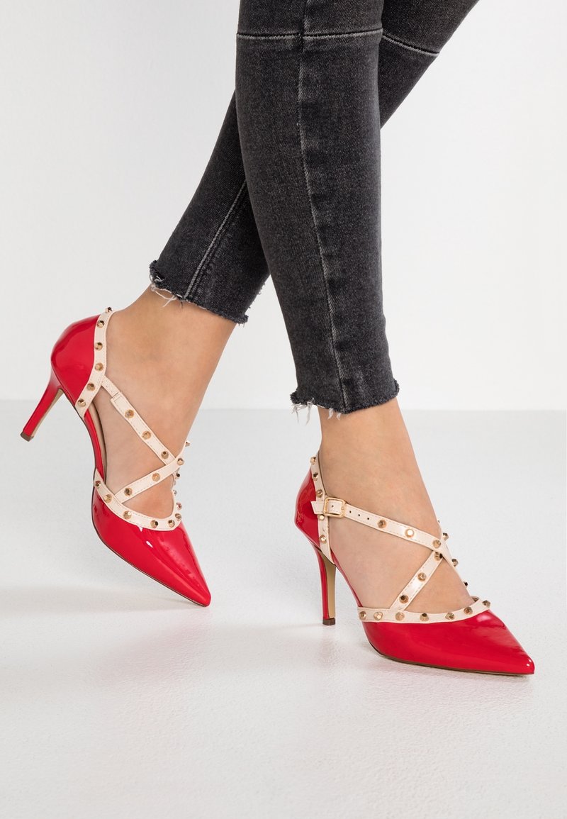 Jambes en jean noir portant des talons aiguilles rouges avec des brides beiges cloutées et des boucles sur fond blanc.