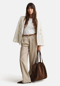 Žena koja nosi bijelu blazu preko bijele košulje i bež hlače na tanke pruge, drži veliki smeđi torbu od brušene kože, stoji na bijeloj pozadini.