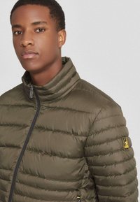 Olijfgroene pufferjas met een hoge kraag, geborduurd ontwerp en zijrits. Heeft een logopatch op de linkermouw. Vlotte textuur.