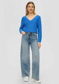 Blauer Strickpullover mit V-Ausschnitt, kombiniert mit hoch taillierten, hellblauen, weit geschnittenen Jeans und weißen Sneakern.
