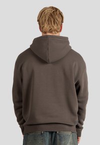 Brauner Kapuzenpullover mit strukturierter Oberfläche, lockerer Passform, geripptem Saum und Manschetten, kombiniert mit verwaschenen Jeans, vor einem hellen Hintergrund präsentiert.