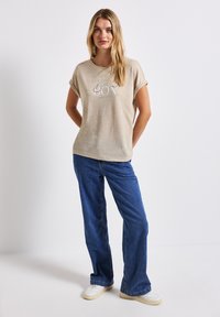 Camisa de manga corta beige con gráfico "BON", combinada con jeans de pierna ancha azules y zapatillas blancas. Diseño simple y casual con ajuste relajado.