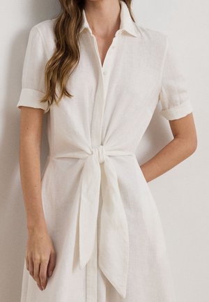 Femme portant une robe blanche à manches courtes avec col et un nœud à l'avant, debout devant un arrière-plan clair uni avec les cheveux sur une épaule.