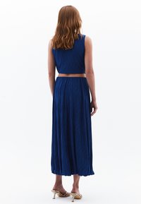 Blauwe twee-delige outfit met een mouwloze top en een lange, vloeiende rok van textuurstof. Het model draagt gouden hoge hakken.