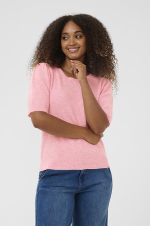 Mujer con cabello rizado que lleva un suéter rosa de manga corta y jeans azules, sonriendo con una mano en la barbilla y la otra cruzada.