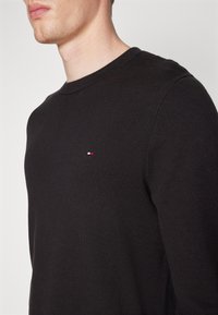 Homme portant un pull ras du cou noir uni avec un petit logo rectangulaire rouge, blanc et bleu sur le côté gauche de la poitrine.