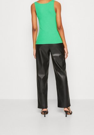 Frau, die ein grünes ärmelloses Oberteil, schwarze Weitbein-Lederhose und schwarze Sandalen mit hohen Absätzen trägt und mit dem Rücken zur Kamera steht.