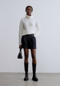 Bela pletena pulover z visokim ovratnikom, temno modri denim kratke hlače, črni škornji do kolen in črna torbica. Gladke teksture in enostavni oblikovni elementi.
