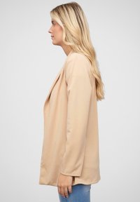 Beige, hosszú ujjú dzseki nyitott elülső résszel, sima textúrával és laza fazonnal, oldalnézetből, sima háttér előtt bemutatva.