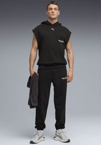 Άνδρας στέκεται, φοράει μαύρο αμάνικο φούτερ με κουκούλα και παντελόνια φόρμας με λογότυπα Puma, κρατώντας μια μαύρη πετσέτα, με λευκά αθλητικά παπούτσια σε γκρι φόντο.