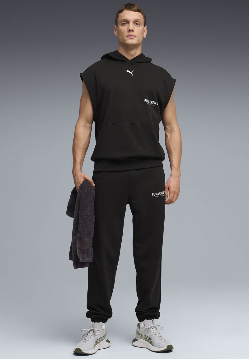 Άνδρας στέκεται, φοράει μαύρο αμάνικο φούτερ με κουκούλα και παντελόνια φόρμας με λογότυπα Puma, κρατώντας μια μαύρη πετσέτα, με λευκά αθλητικά παπούτσια σε γκρι φόντο.