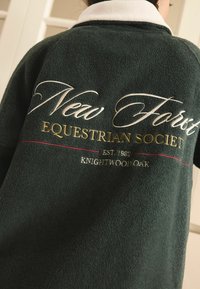 Vihreä fleece-takki valkoisella kauluksella, jossa on kirjailtuna teksti: "New Forest Equestrian Society, EST. 1982, Knightwood Oak."