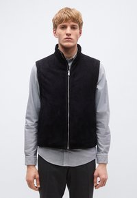 Fekete ujjatlan mellény sima anyagból, magas gallérral és elöl cipzárral, szürke, gombos ing fölött viselve. Testhezálló szabás.