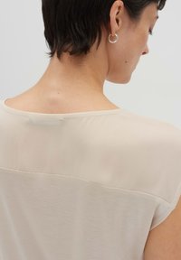 Beige Bluse mit einem transparenten oberen Abschnitt und einem soliden unteren Teil. Das Design umfasst kurze Ärmel und eine glatte Textur. Mit sauberen Nähten akzentuiert.