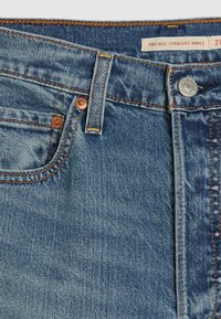 Zone avant de la taille d'un jean en denim bleu avec surpiqûres orange, bouton en métal, rivets, passants de ceinture et étiquette portant la mention « Ribcage Straight Ankle ».