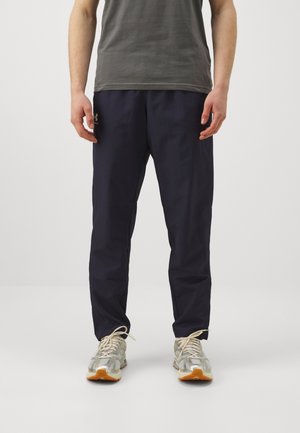 Mężczyzna ubrany w ciemnoniebieskie spodnie, szare buty sportowe z białymi sznurowadłami oraz ciemnoszarą koszulkę, stojący na jednolitym jasnym tle.