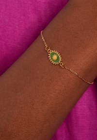 Gouden armband met een ovale groene centraal stuk met een zonontwerp, gedragen op een donkere huidarm tegen een fuchsia achtergrond.