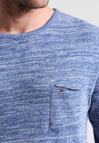 Chemise à manches longues en tricot bleu avec une finition texturée, dotée d'une petite poche poitrine. La poche arbore un logo distinctif en rouge, blanc et bleu.