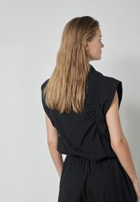 Čierny tielkový top s voľným strihom, s nariasenými detailmi na zadnej strane a hladkou textúrou. Modelka má dlhé rovné vlasy.