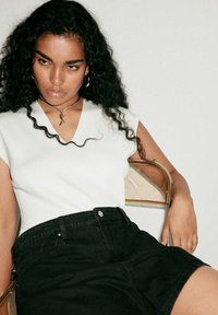 Jeune femme aux longs cheveux bouclés portant un haut blanc sans manches et un short noir, assise sur une chaise en métal contre un mur uni.