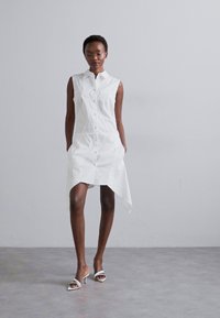 Fehér ujjatlan ingruha gombos elülső résszel, aszimmetrikus szegéllyel és sima pamut anyaggal. Fehér magassarkú szandálokkal viselve.