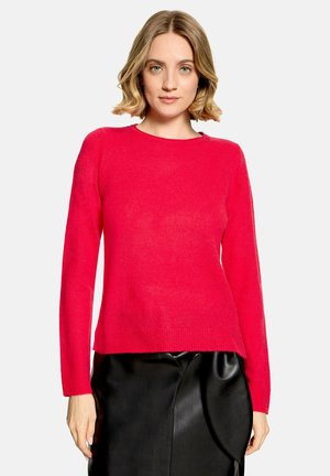Femme aux cheveux blonds courts portant un pull rouge vif à manches longues et une jupe en cuir noire sur un fond uni.