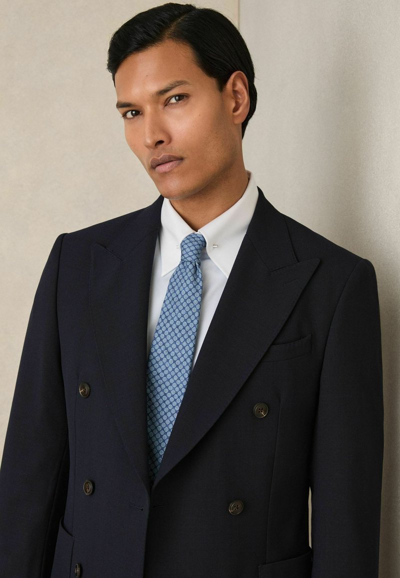 Dubbelzijdig marineblauwe blazer met bruine knopen, gecombineerd met een lichtblauwe gestippelde stropdas en een witte overhemden. Zachte textuur en nauwsluitende pasvorm.