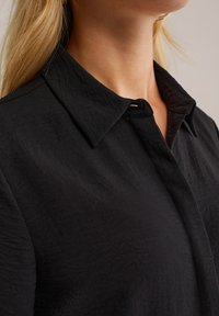 Donna con lunghi capelli biondi che indossa una camicia nera con colletto, ripresa dalla zona del collo al petto su uno sfondo neutro.