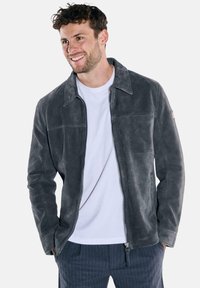 Graue Lederjacke mit Reißverschluss vorne, breitem Kragen und einer Brusttasche. Glatte Textur mit dezentem Glanz, getragen über einem weißen Hemd.