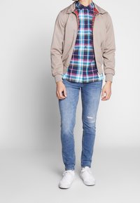 Veste bomber légère beige avec des poignets élastiques, superposée à une chemise à carreaux multicolores avec des carrés bleus, rouges et blancs. Portée avec un jean bleu déchiré et des baskets blanches.