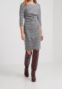 Robe grise moulante avec un motif à carreaux diagonal, manches trois-quarts et un détail torsadé à la taille, associée à des bottes bordeaux montant jusqu'au genou.