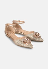Ballerines dorées avec brides au niveau de la cheville, présentant un dessus texturé et un ornement bijou sur le bout. Semelle plate, hauteur minimale.