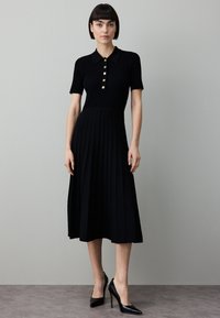 Svart ribbad klänning med korta ärmar, krage med knappar och plisserad kjol. Matchad med spetsiga svarta klackskor mot en neutral bakgrund.