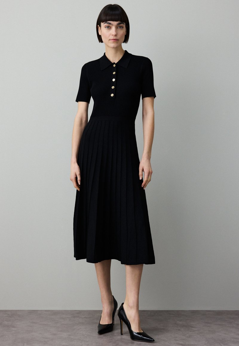 Svart ribbad klänning med korta ärmar, krage med knappar och plisserad kjol. Matchad med spetsiga svarta klackskor mot en neutral bakgrund.