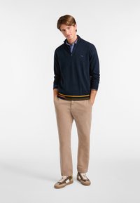 Marineblauer Zip-Kragen-Pullover mit gestreiftem Saum, beigen Chinos und braunen Sneakers mit grünen und gelben Akzenten. Glatte Textur, lässiger Schnitt.