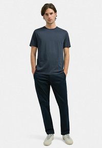 Mörkgrå t-shirt med kort ärm och rund halsringning, tillsammans med åtsittande marinblå byxor. Modellen bär ljusa sneakers och står avslappnat.